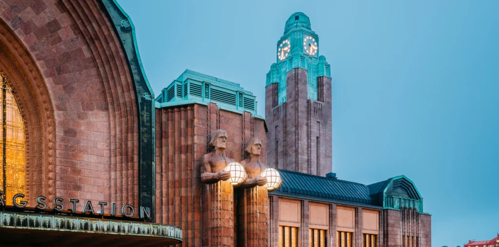 Close-up of Helsinki Central Railway Station's Art Nouveau facade at dusk, with two illuminated statues holding glowing globe lamps beneath the tall green-domed clock tower and warm light glowing from the arched entrance.