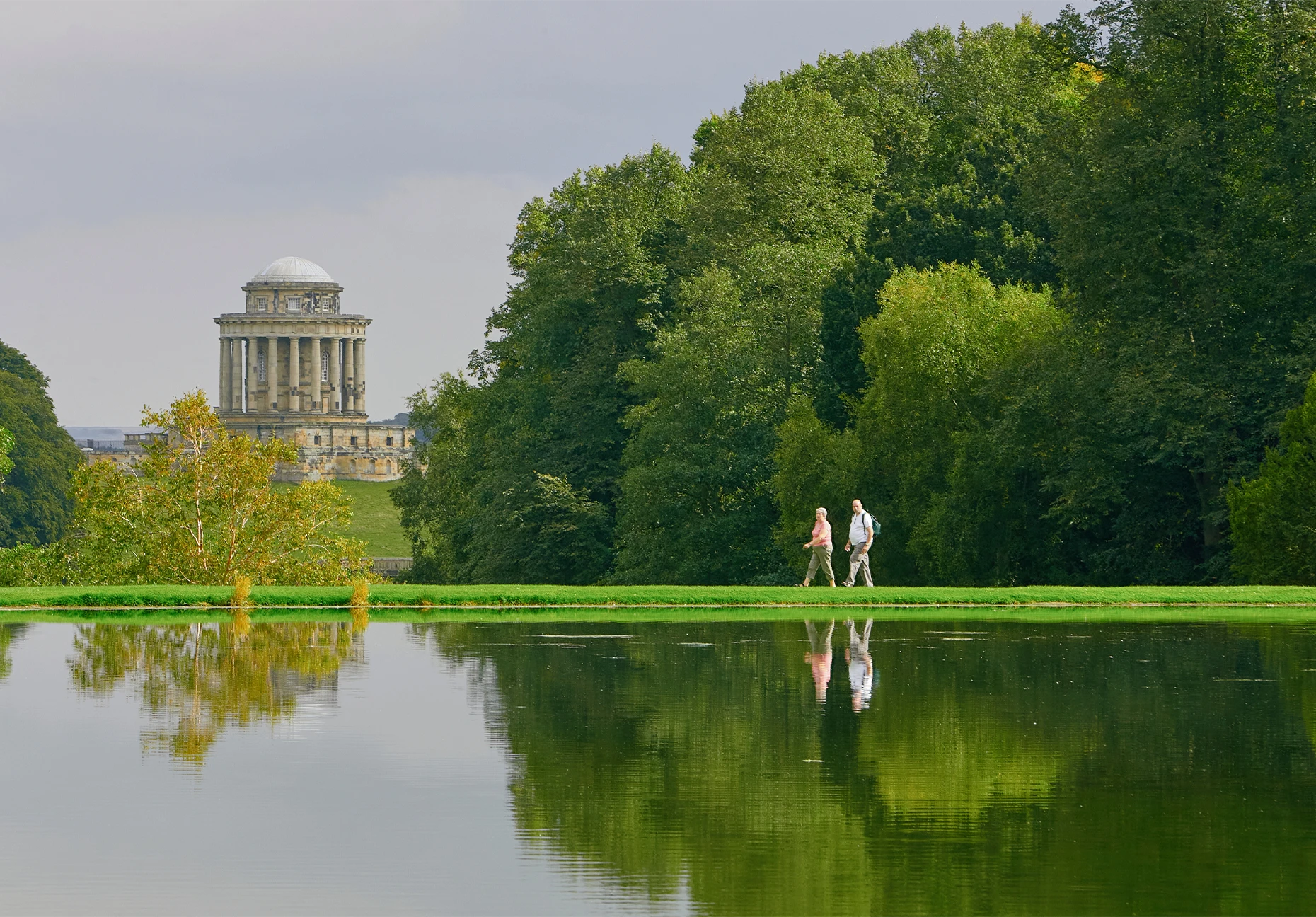 Two walkers pass a still lake with a domed classical mausoleum beyond the trees
