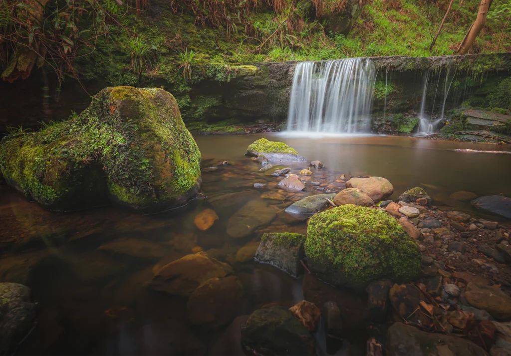 A wide waterfall spills over a mossy ledge into a shallow pool ringed by boulders