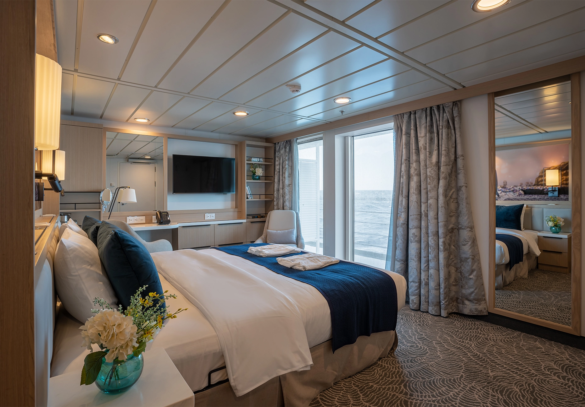 Bed with navy runner and folded robes beside balcony doors and a wall-mounted television