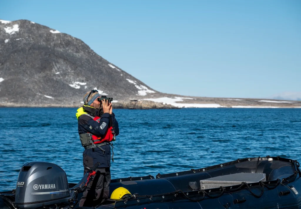 Man with binoculars standing in a Zodiac beside a dark scree slope and blue water
