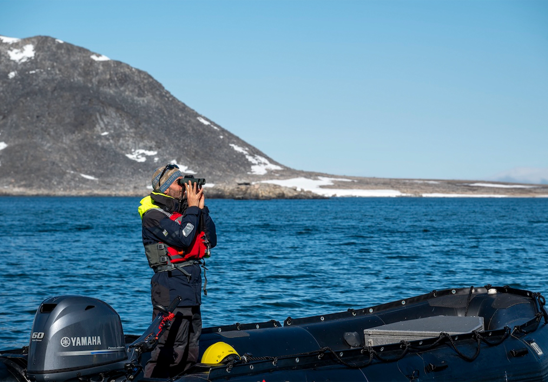 Man with binoculars standing in a Zodiac beside a dark scree slope and blue water