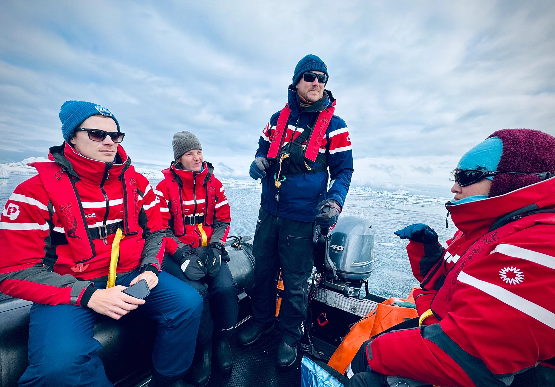 Four people in red expedition jackets aboard an inflatable boat surrounded by floating ice