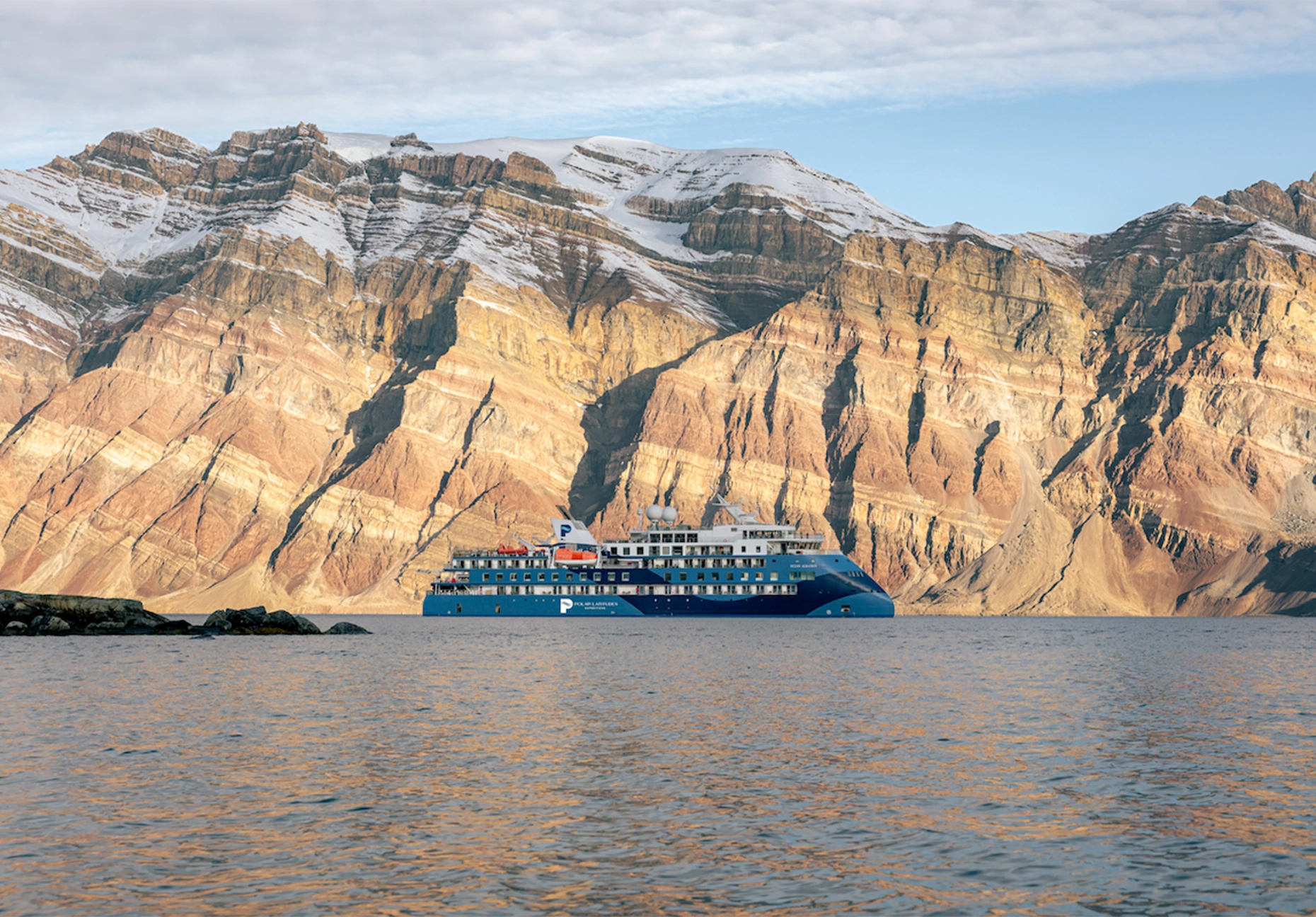 Blue expedition vessel at anchor beneath towering banded rock cliffs topped with snow