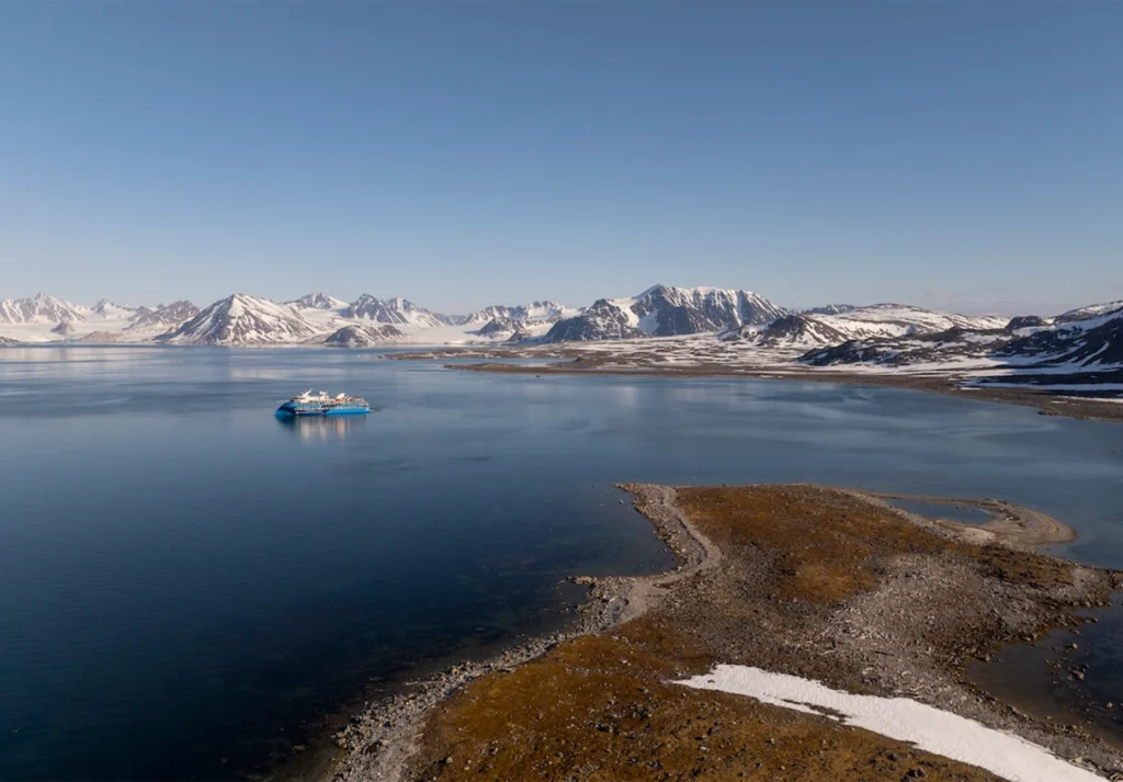 Small blue expedition vessel on calm water below snow-covered mountains and a low tundra spit