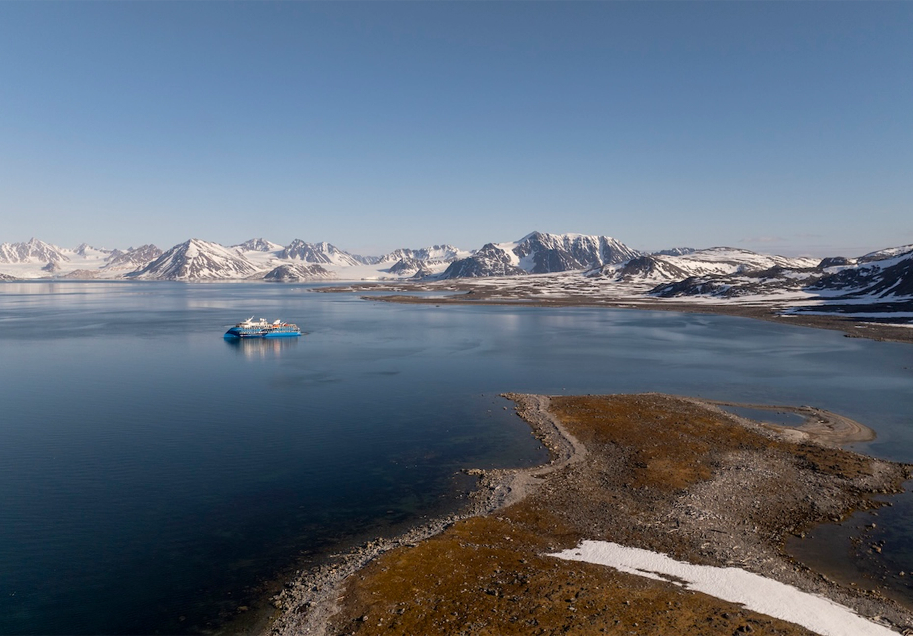 Small blue expedition vessel on calm water below snow-covered mountains and a low tundra spit