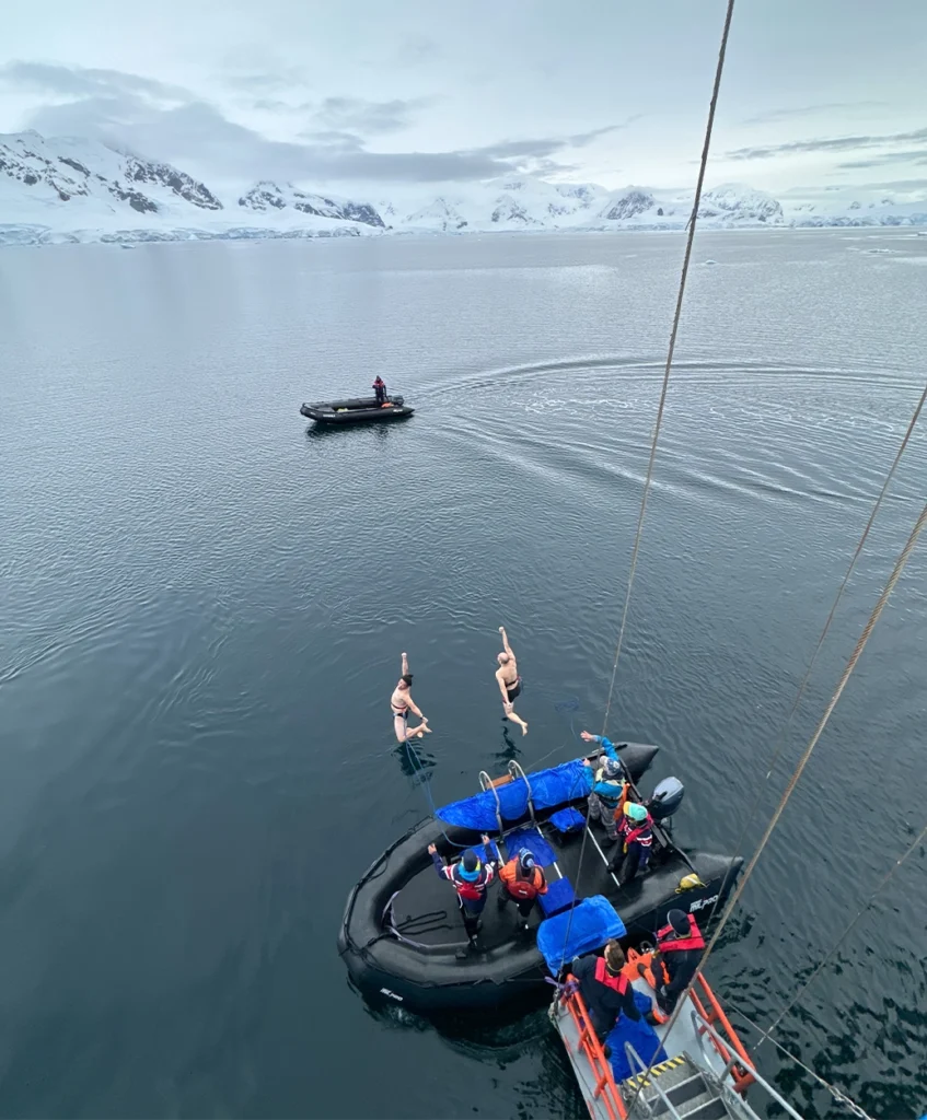 Two swimmers leaping into icy water beside Zodiacs with snow-covered peaks behind