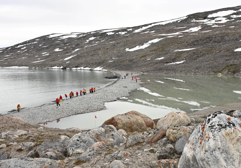 Line of people in red jackets crossing a stony spit beside a Zodiac and still water