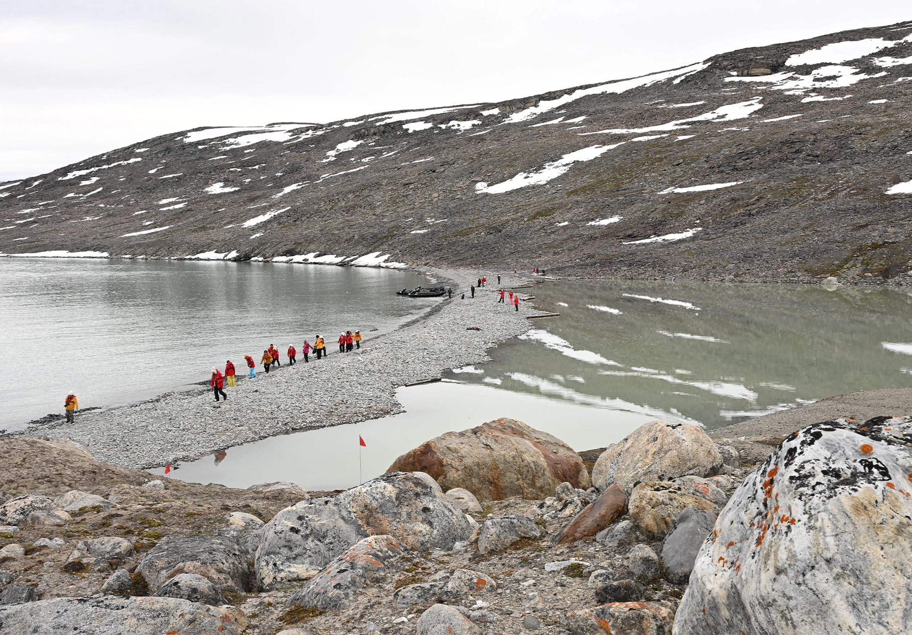 Line of people in red jackets crossing a stony spit beside a Zodiac and still water