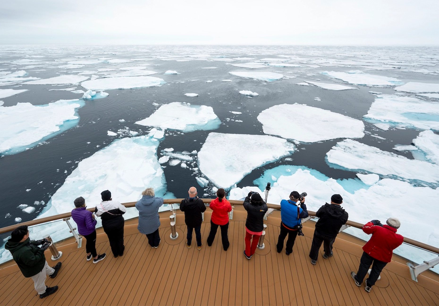 Row of passengers at a curved deck railing overlooking scattered white ice floes