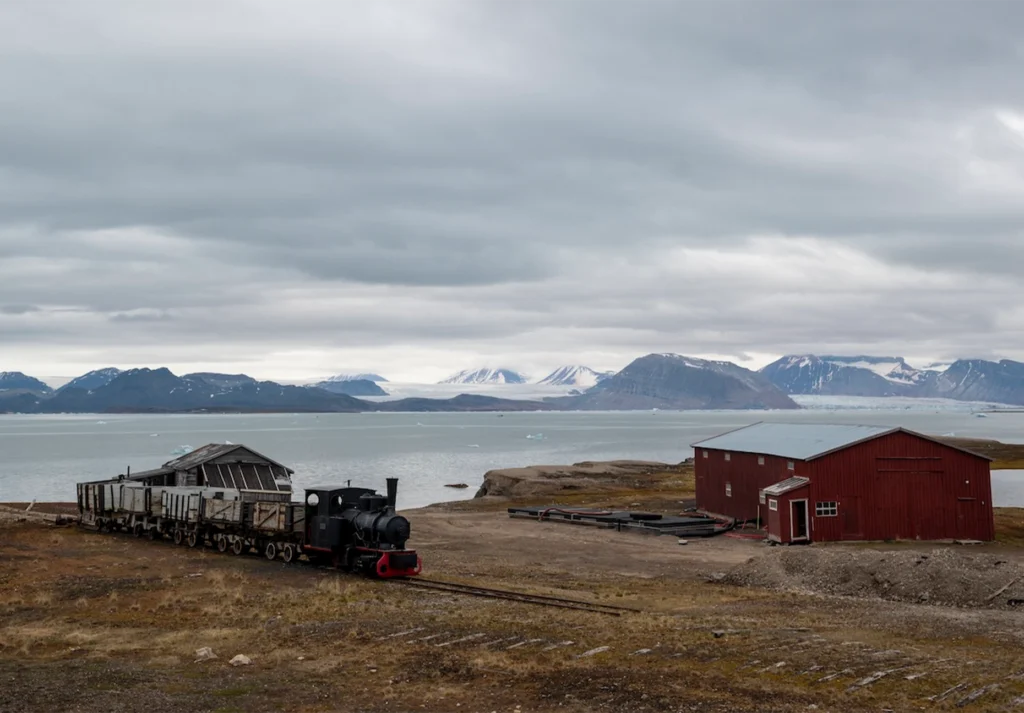 Preserved steam locomotive with ore wagons beside a red barn and glacier-backed fjord