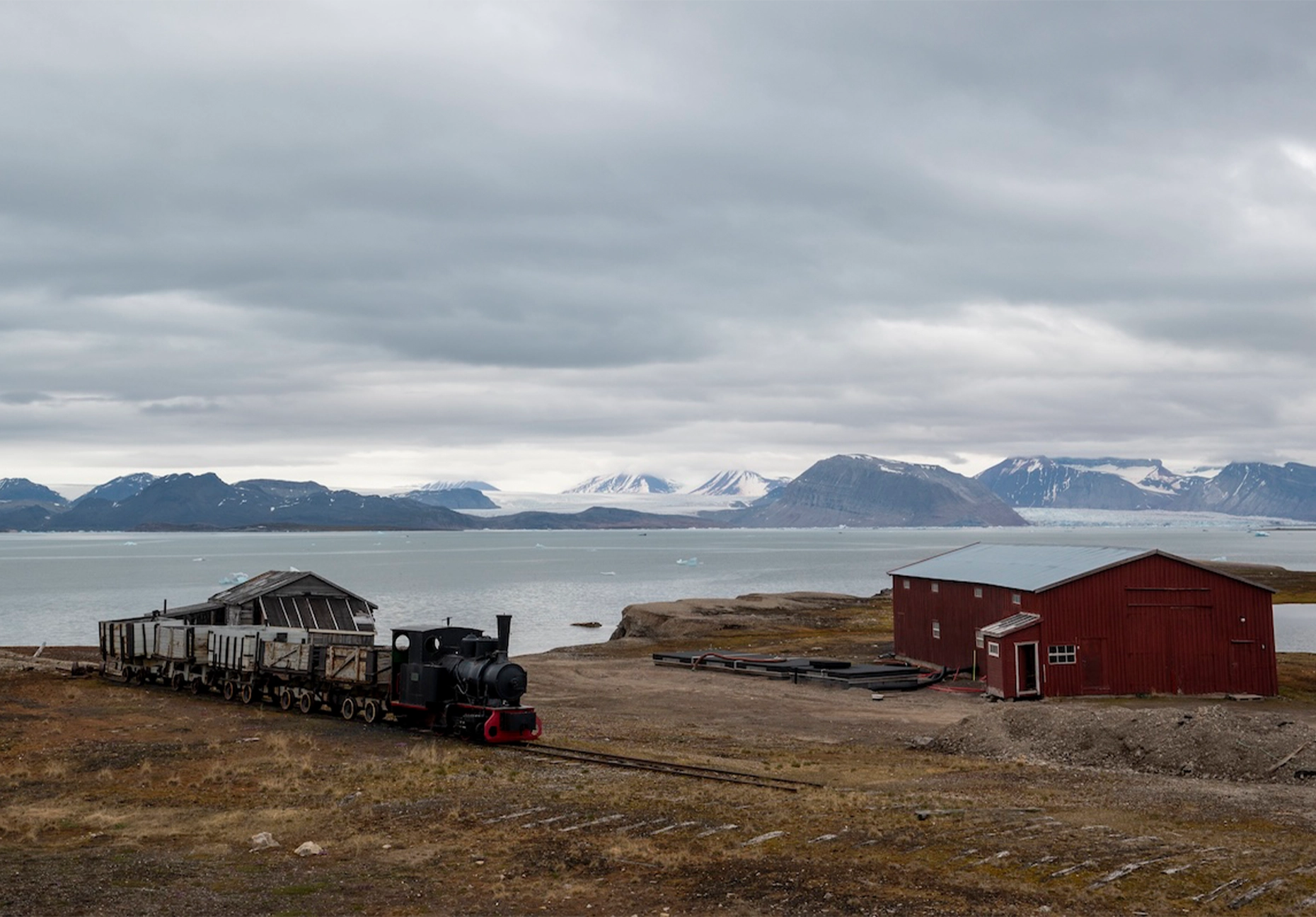 Preserved steam locomotive with ore wagons beside a red barn and glacier-backed fjord