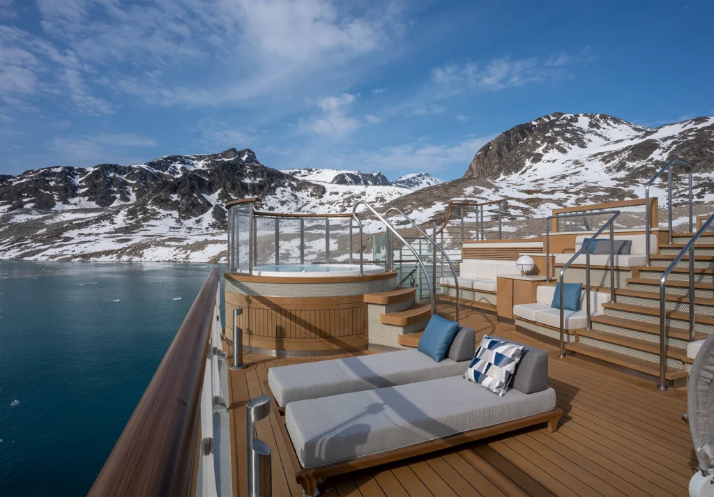 Circular hot tub and cushioned loungers on a teak deck facing snowy peaks