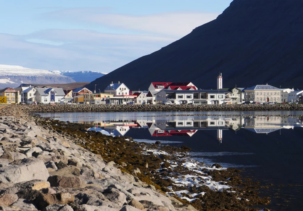 Coastal town buildings mirrored in calm fjord water beneath a steep dark mountain