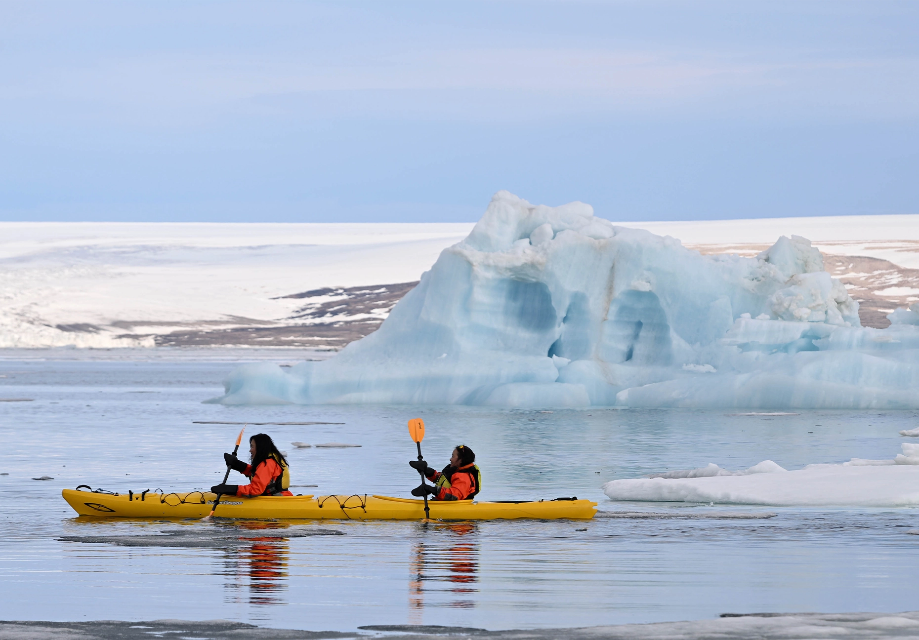 Two people in orange drysuits in a yellow kayak beside a pale blue iceberg