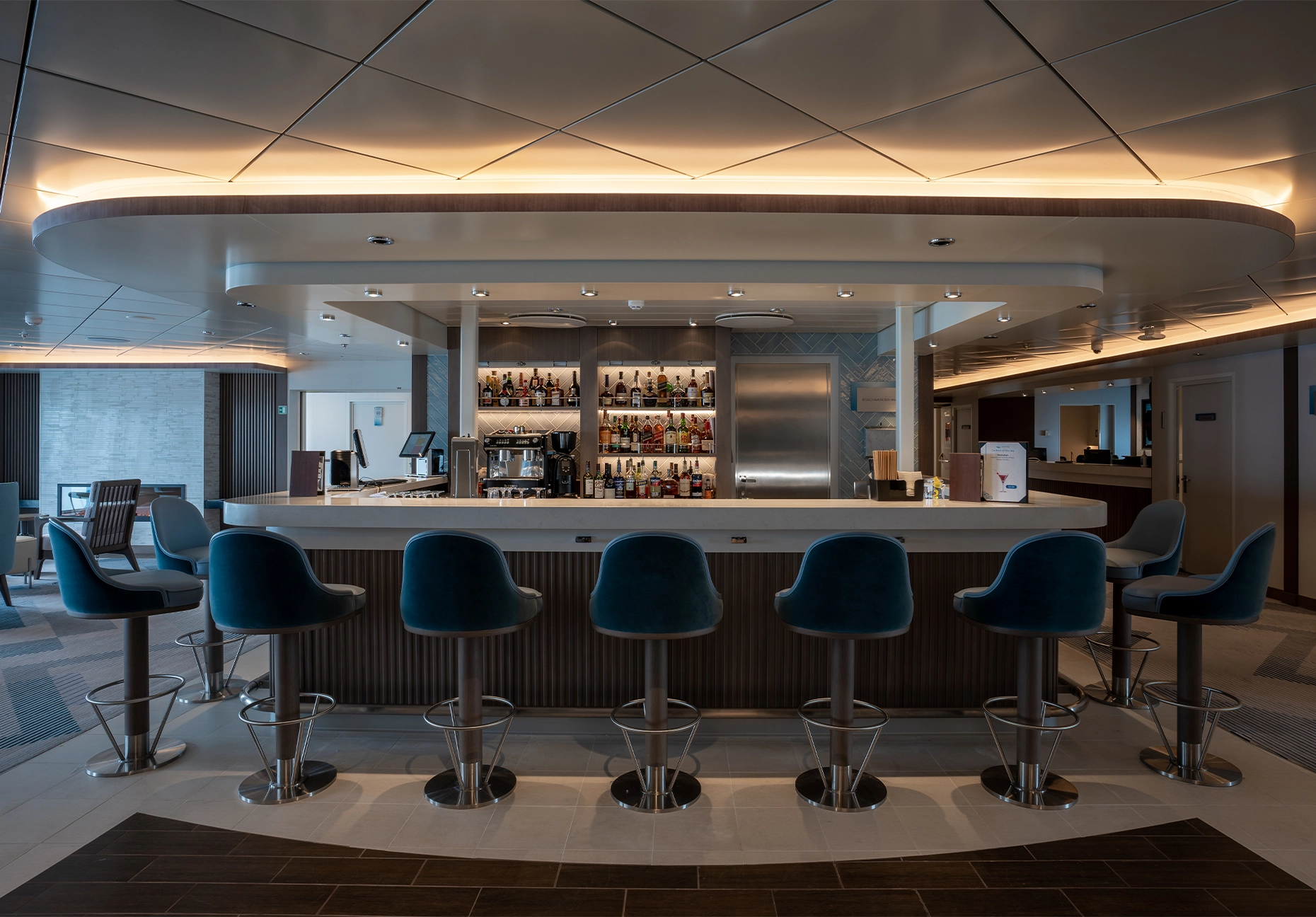 Curved bar with teal velvet stools and backlit bottle shelves under warm cove lighting