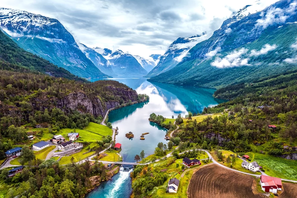 Aerial view of a dramatic Norwegian fjord landscape with a still turquoise lake reflecting snow-capped mountains and clouds, flanked by steep forested valley walls. Traditional red and white farmhouses and winding rural roads dot the lush green valley floor, with a rushing glacial river flowing beneath a small bridge in the foreground.
