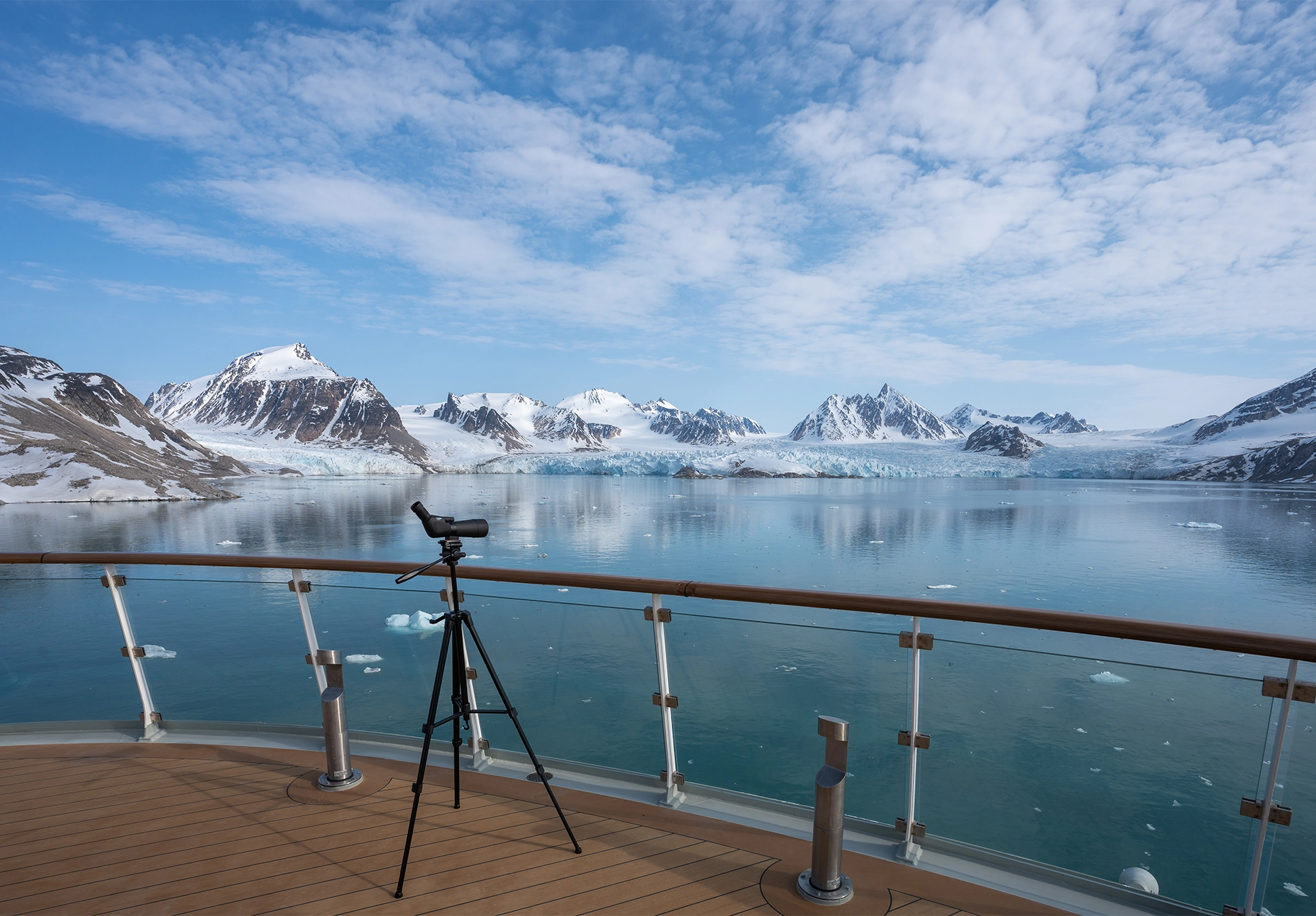 Spotting scope on a teak deck facing a glacier, ice-strewn water, and snow-capped peaks