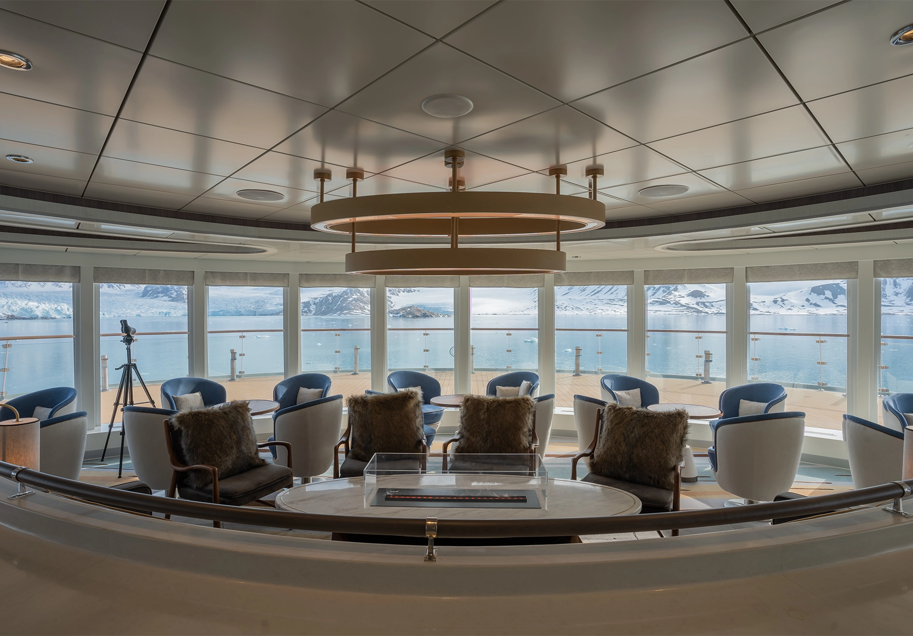 Armchairs with fur cushions around a fire table facing curved windows over a glacier bay