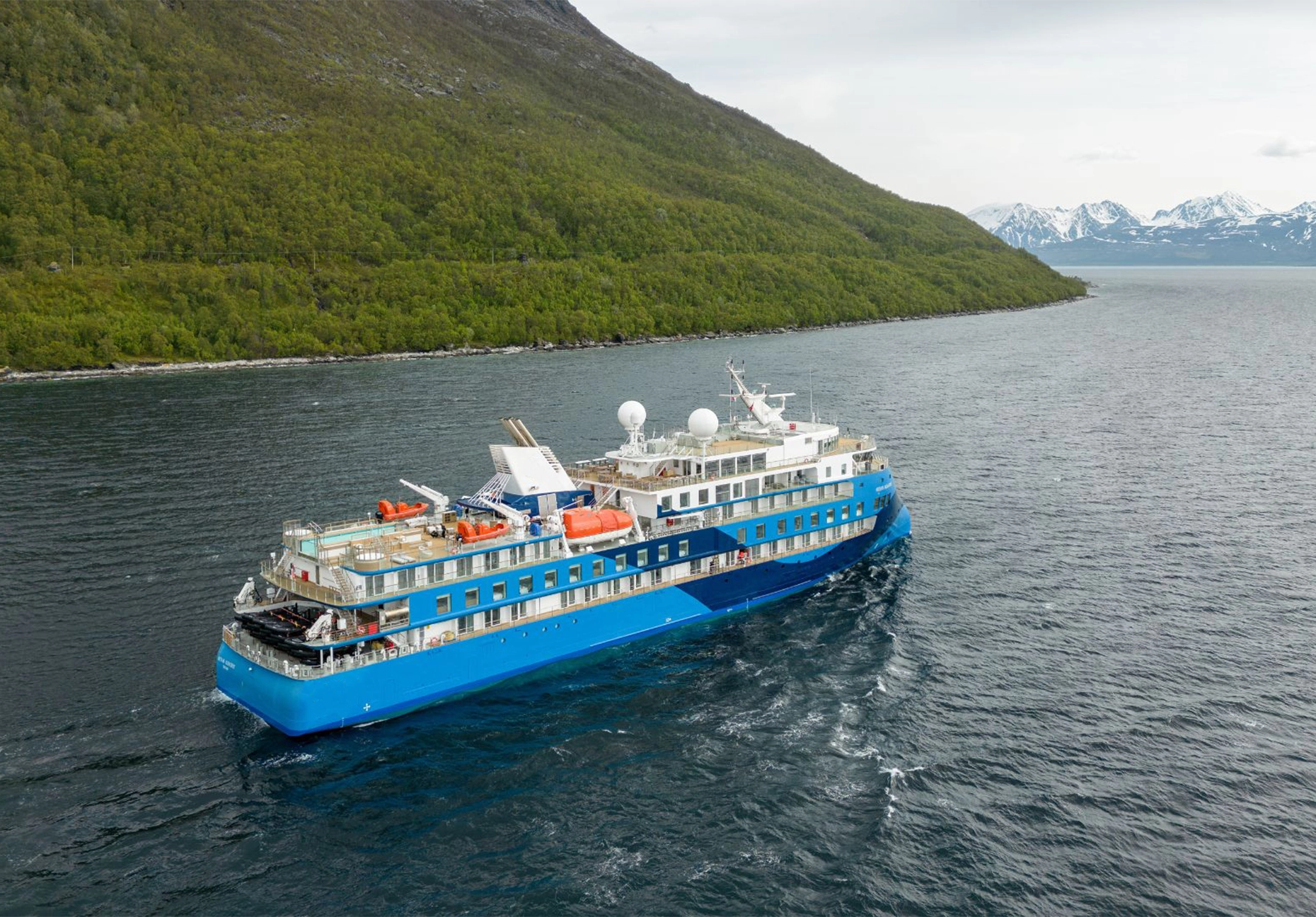 Blue and white expedition ship cruising past a wooded hillside with distant snowy mountains