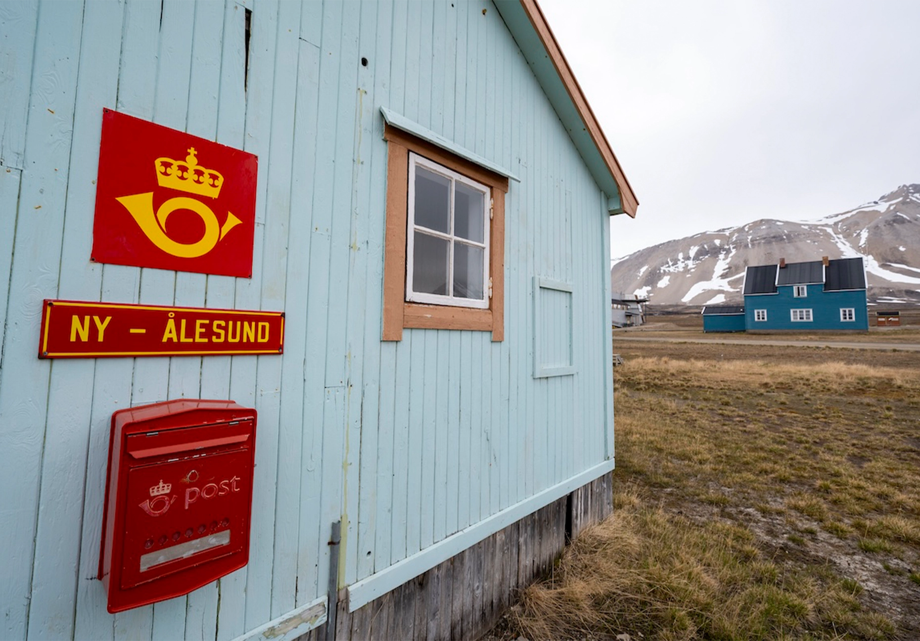 Red Norwegian post sign and mailbox on a pale blue timber wall with mountains behind