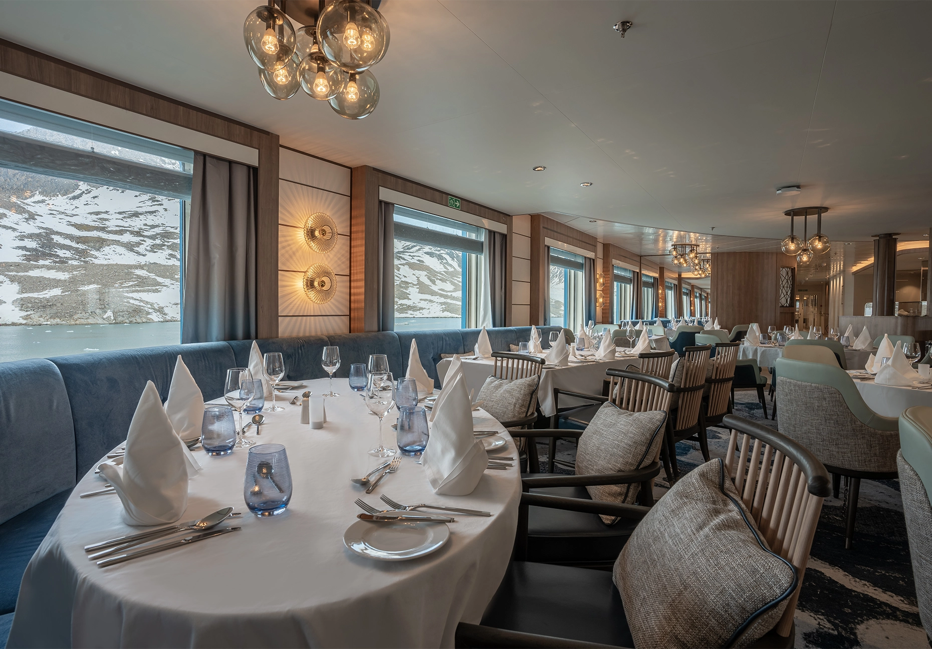 Set dining tables with white linen and blue velvet banquettes beside snow-covered mountain views