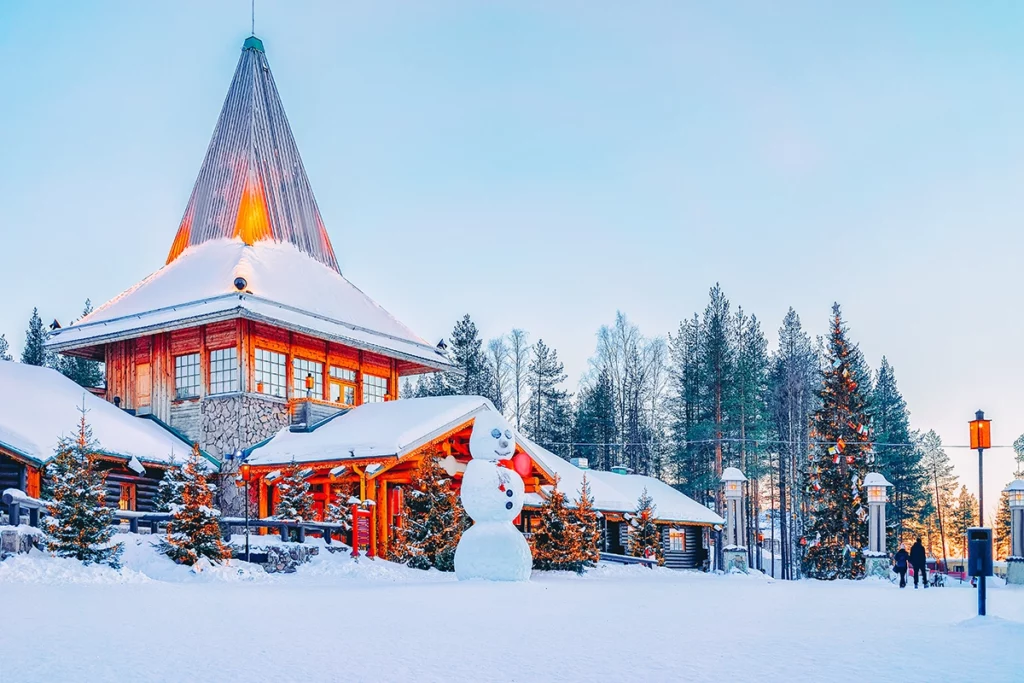 Santa Claus Village in Rovaniemi, Finland, in winter, featuring a large snow-covered log cabin with a tall pointed metal roof glowing with warm light. A large snowman stands in the foreground, surrounded by snow-dusted Christmas trees strung with lights, pine forests, and vintage street lamps under a soft pastel winter sky