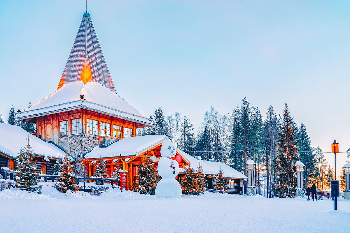 Santa Claus Village in Rovaniemi, Finland, in winter, featuring a large snow-covered log cabin with a tall pointed metal roof glowing with warm light. A large snowman stands in the foreground, surrounded by snow-dusted Christmas trees strung with lights, pine forests, and vintage street lamps under a soft pastel winter sky
