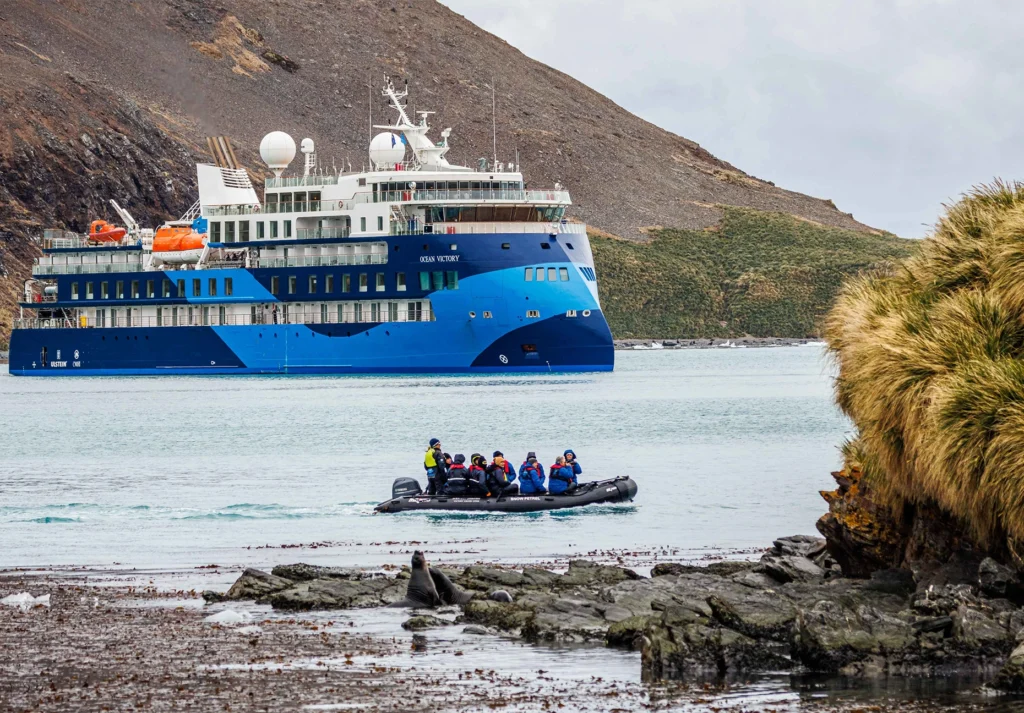 Blue expedition ship anchored offshore as passengers ride an inflatable boat past a rocky coast