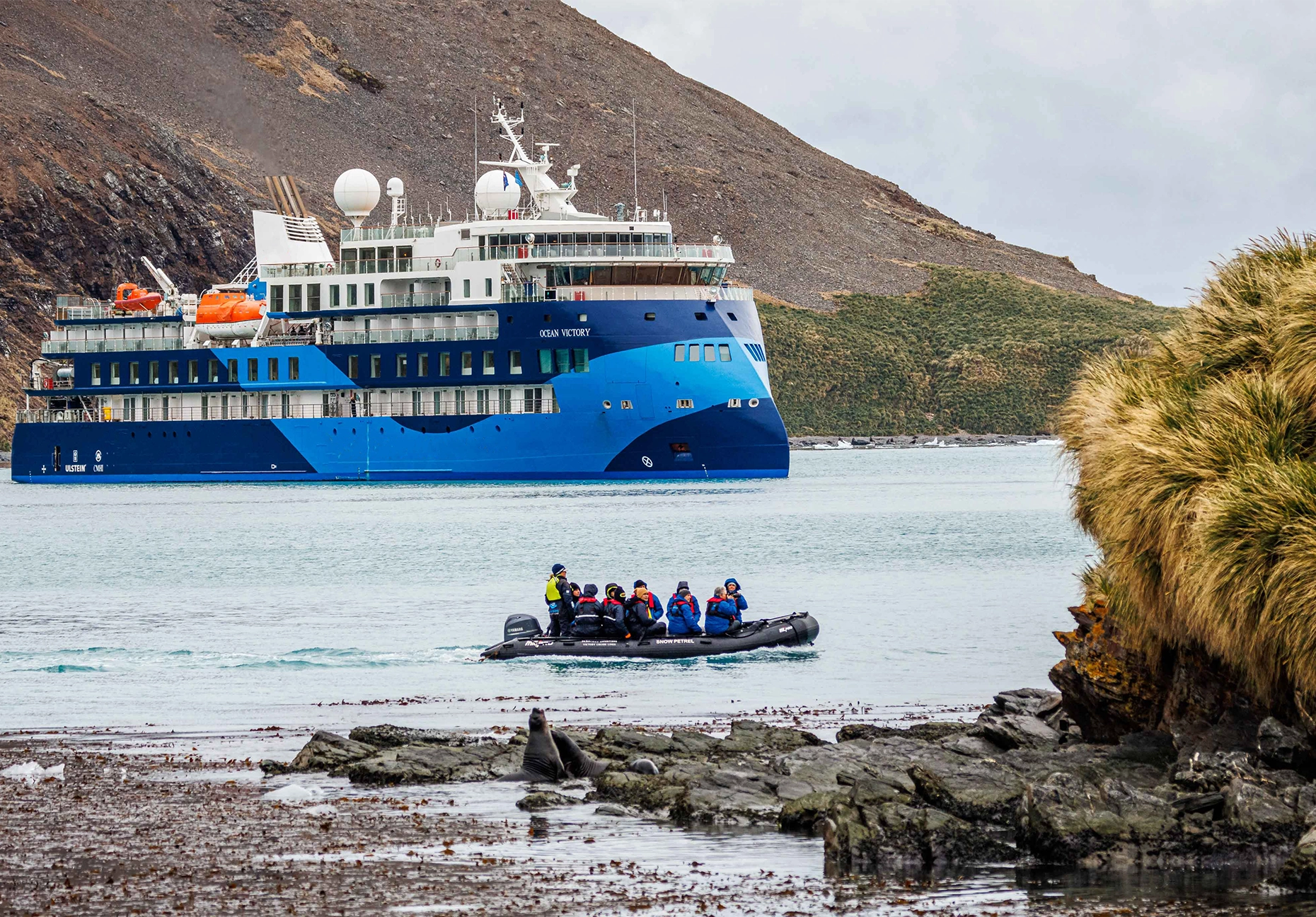 Blue expedition ship anchored offshore as passengers ride an inflatable boat past a rocky coast