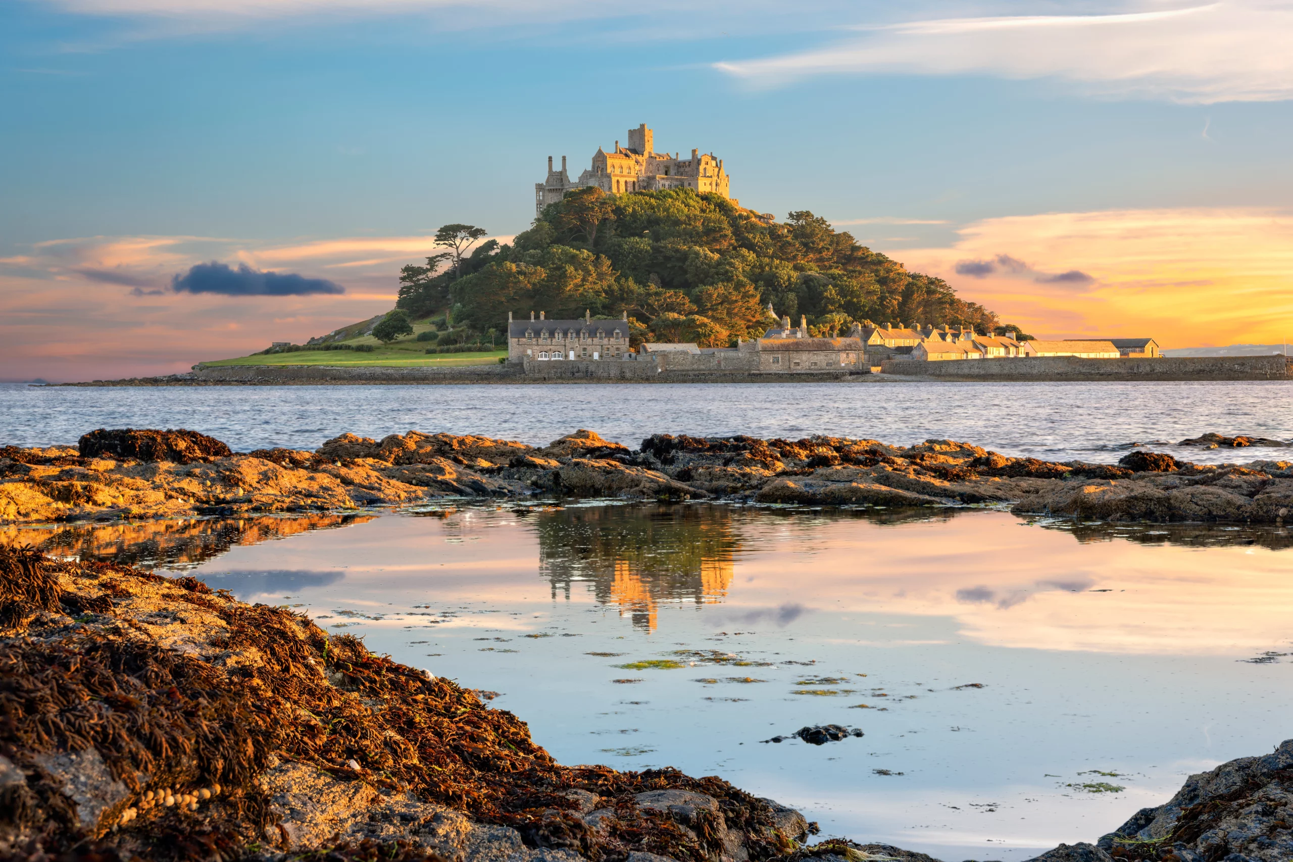 St Michael's Mount, a medieval castle and chapel perched atop a tree-covered tidal island in Cornwall, England, photographed at sunset. The golden-lit castle and surrounding stone harbour buildings are reflected in a shallow coastal rock pool in the foreground, beneath a dramatic sky of orange, pink, and blue.