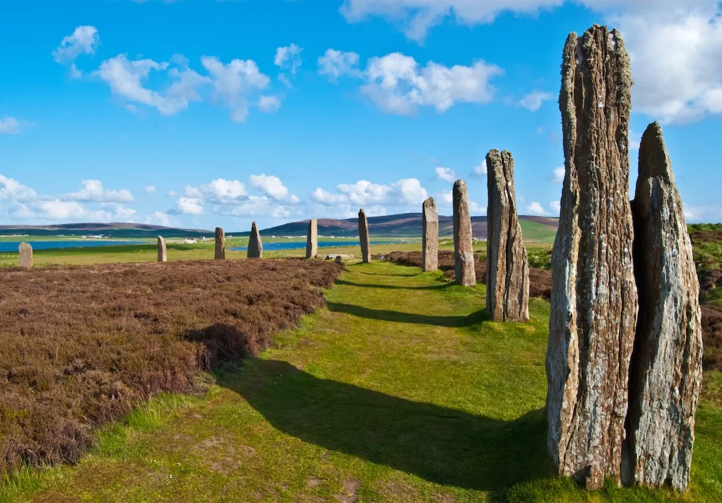 Weathered stone monoliths in a grassy circle with heather and a loch behind