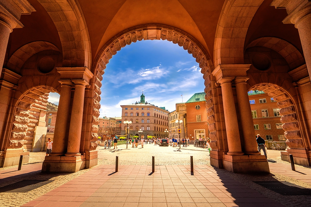 View through a grand sandstone archway into a sun-drenched public square in Stockholm, Sweden, framed by ornate red-stone columns and vaulted ceilings. Beyond the arch, pedestrians walk across a cobblestone plaza lined with warm-toned historic buildings, one topped with a green copper spire, under a bright blue sky
