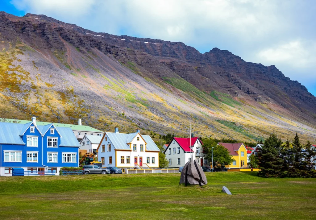Brightly painted wooden houses on a green lawn below a steep scree-covered mountain