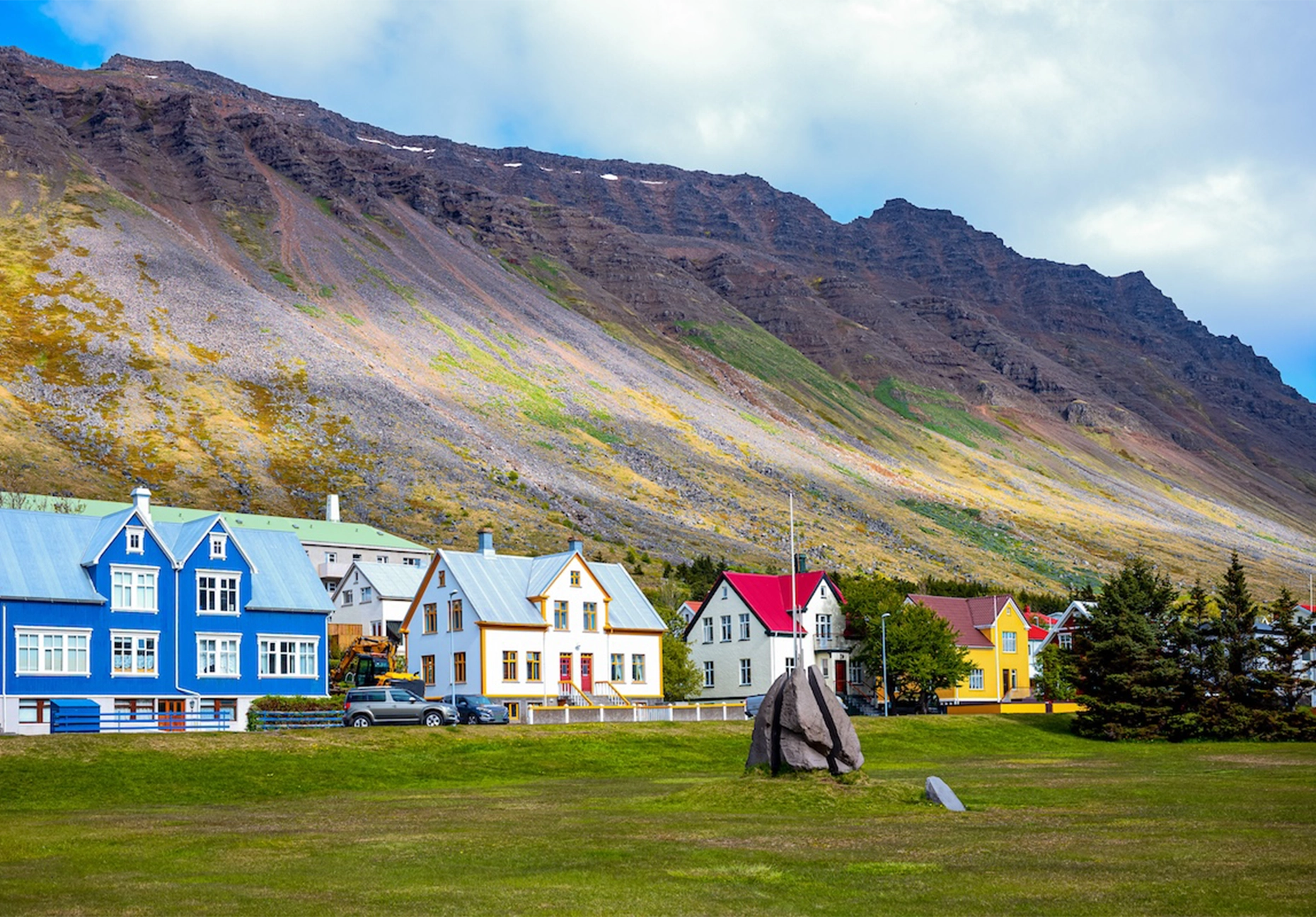 Brightly painted wooden houses on a green lawn below a steep scree-covered mountain