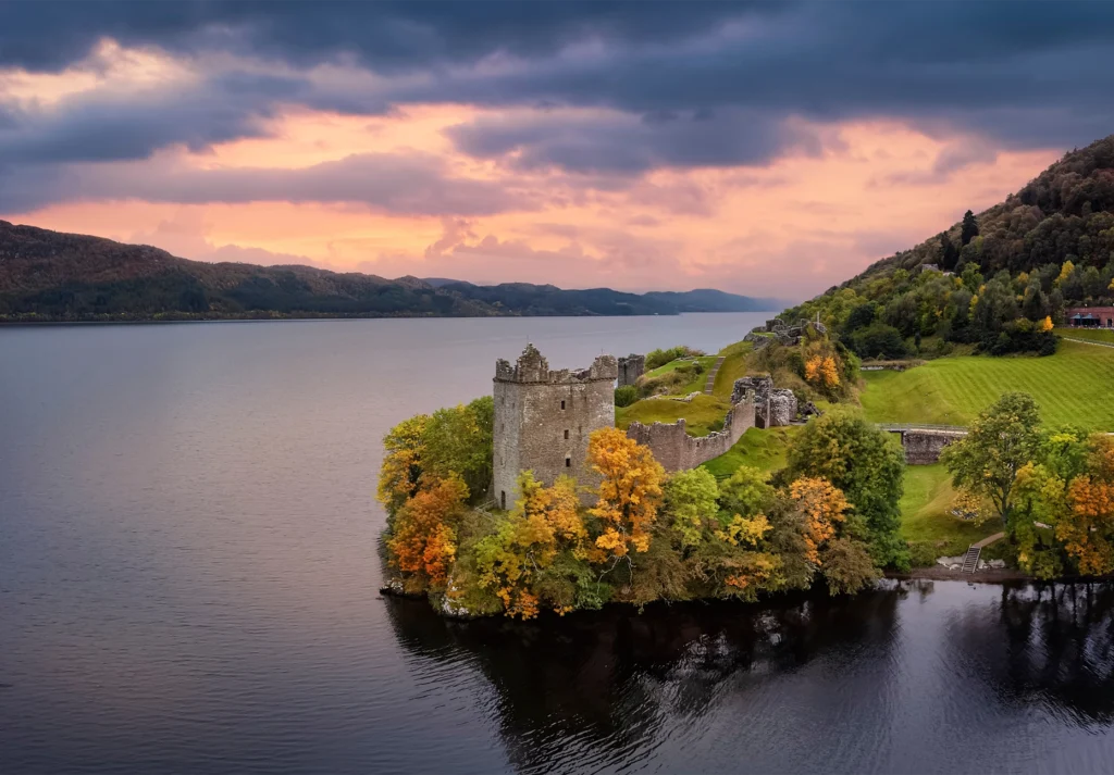 Ruined stone tower among autumn trees beside dark loch water under a pink sky