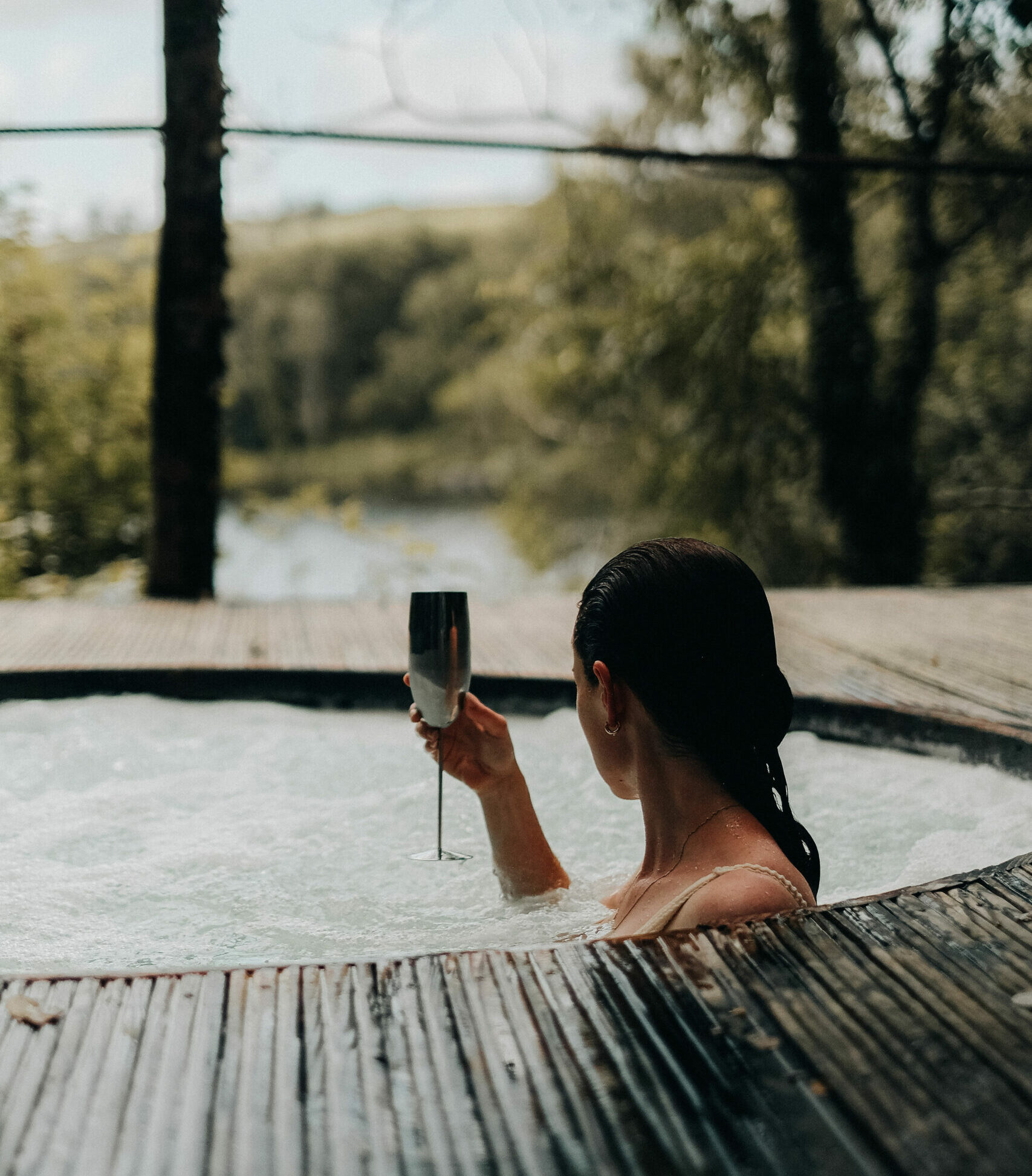 A woman relaxes in an outdoor wooden hot tub holding a champagne flute at a wellness retreat in Ireland, surrounded by a timber deck and lush green woodland with a tranquil lake visible in the background.