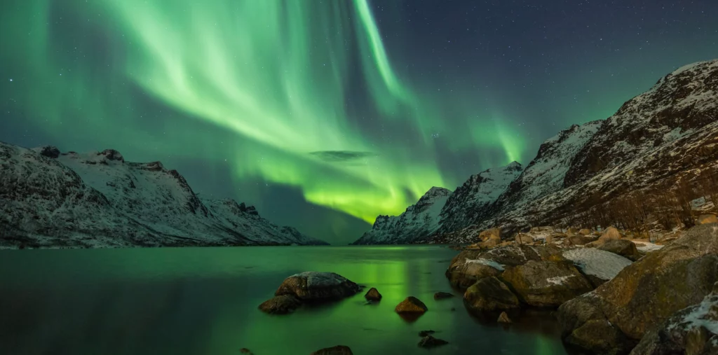 Vivid green northern lights swirling across the night sky above a fjord near Tromsø, Norway, with snow-covered mountains and the aurora reflected in the still water