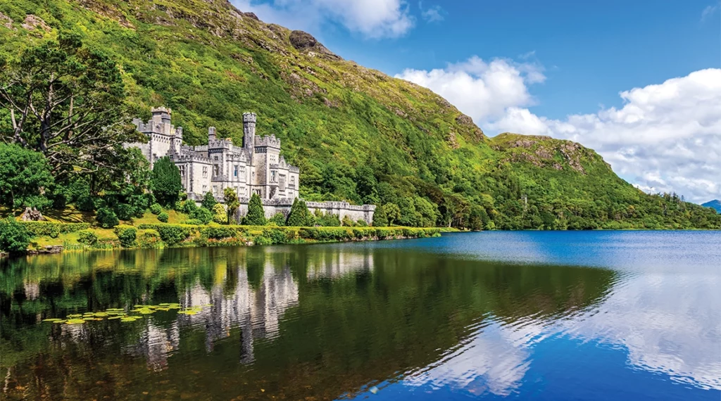 Kylemore Abbey, a grey Gothic castle set against a green wooded mountainside in Connemara, reflected in the still waters of the lake below.