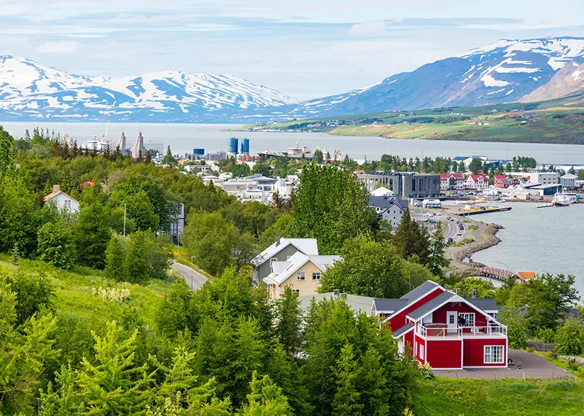 View over Akureyri town with colourful houses, green trees, and the harbour on Eyjafjörður fjord with snow-capped mountains in the background, northern Iceland