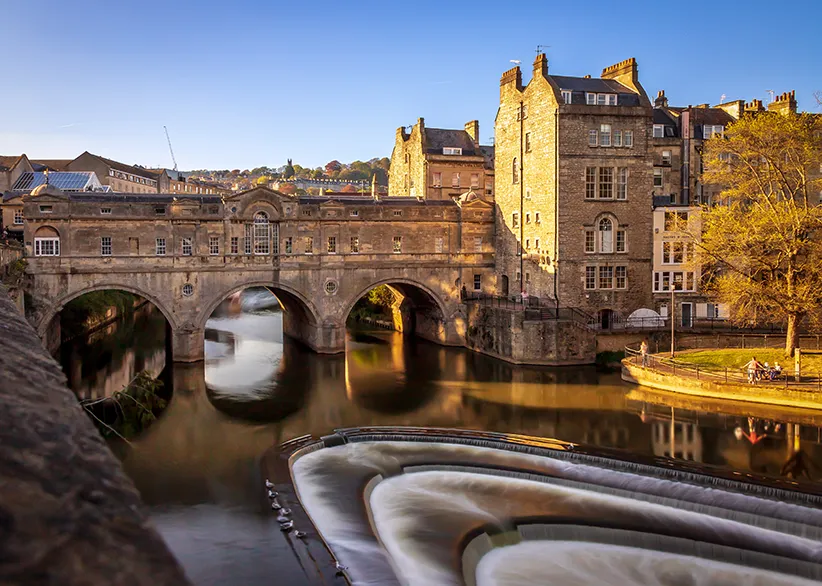 Pulteney Bridge spanning the River Avon with its horseshoe weir in the foreground and honey-coloured Georgian stone buildings in warm evening light, Bath, England