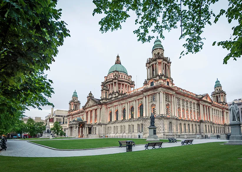 Belfast City Hall's grand Edwardian Baroque facade with green copper domes, framed by trees, with manicured lawns and benches in the foreground