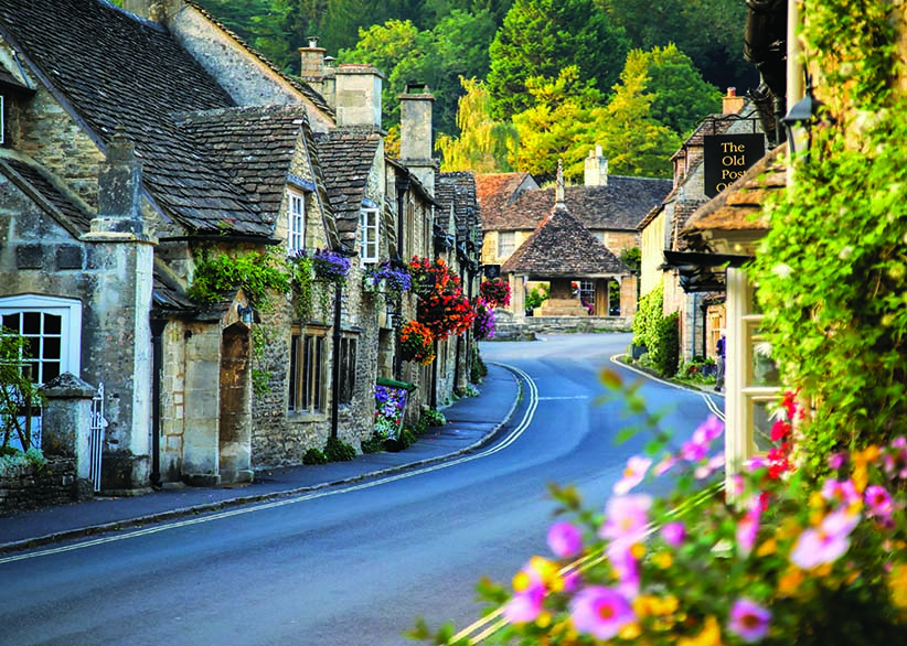 The picturesque main street of Castle Combe village in the Cotswolds with honey-coloured stone cottages, hanging flower baskets, and lush greenery on a summer evening