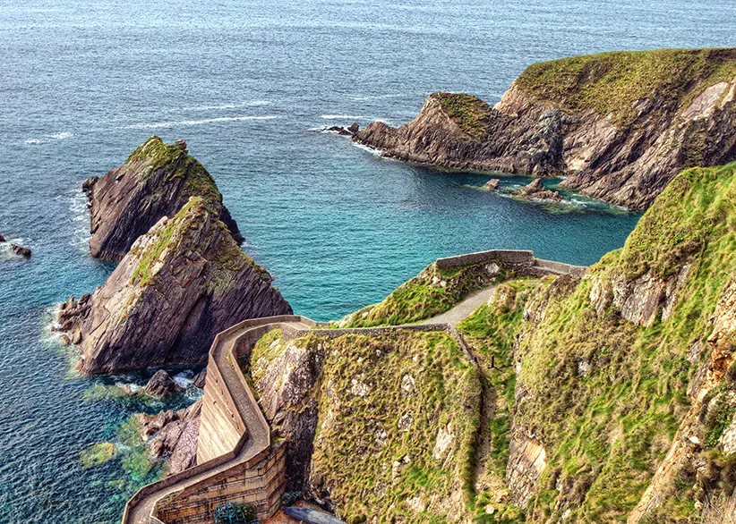 The steep winding path of Dunquin Pier descending through green cliffs to the turquoise Atlantic waters with rugged headlands on the Dingle Peninsula, County Kerry, Ireland