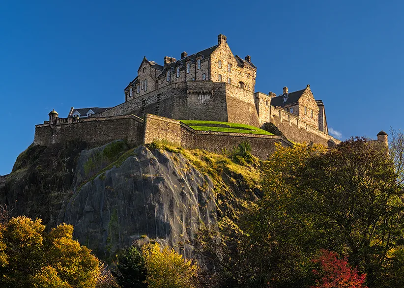 Alt text: Edinburgh Castle perched on its volcanic rock seen from below on a clear autumn day with golden and red foliage framing the castle walls against a deep blue sky