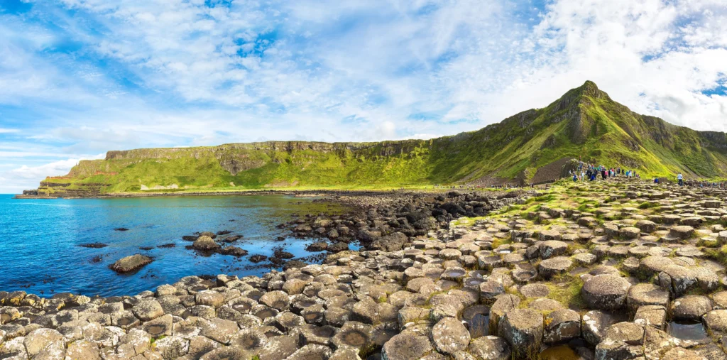 Panoramic view of the Giant's Causeway hexagonal basalt columns stretching towards the sea with visitors exploring, green cliffs, and Aird Snout headland under a blue sky, County Antrim, Northern Ireland