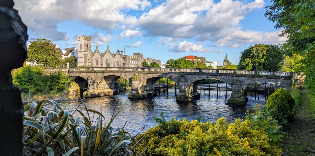 A stone bridge over the River Corrib in Galway city with Galway Cathedral visible in the background, lush greenery in the foreground, and a sunny blue sky