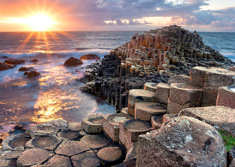 The hexagonal basalt columns of the Giant's Causeway at sunset with waves washing over the rocks and a golden sunburst on the horizon, County Antrim, Northern Ireland
