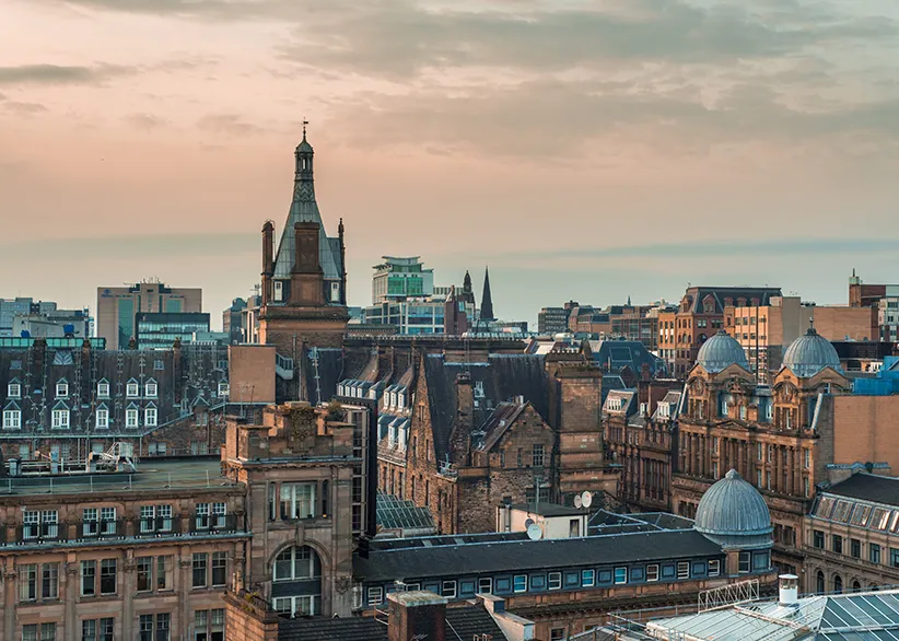 Rooftop view of Glasgow's Victorian and Edwardian sandstone architecture with ornate towers, domes, and spires under a soft pink evening sky