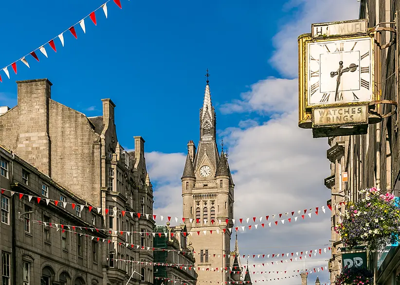Aberdeen's granite buildings and clock tower on a sunny day with red and white bunting strung across the street and a vintage shop clock in the foreground